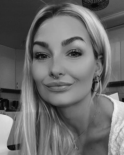 En kvinna med långt blont hår och en septumpiercing ler svagt i ett kök. Bilden är i svartvitt.