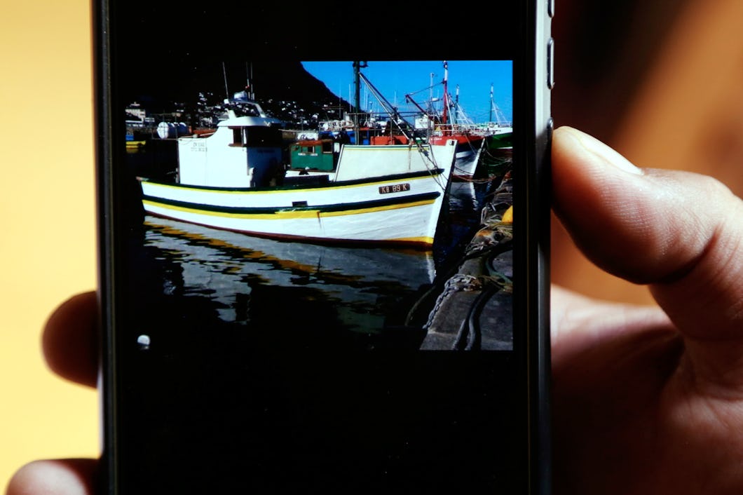 En hand håller en mobiltelefon som visar en bild av en fiskebåt vid kaj med färgerna vitt, gult och grönt. I bakgrunden syns fler båtar och en del av en hamn.