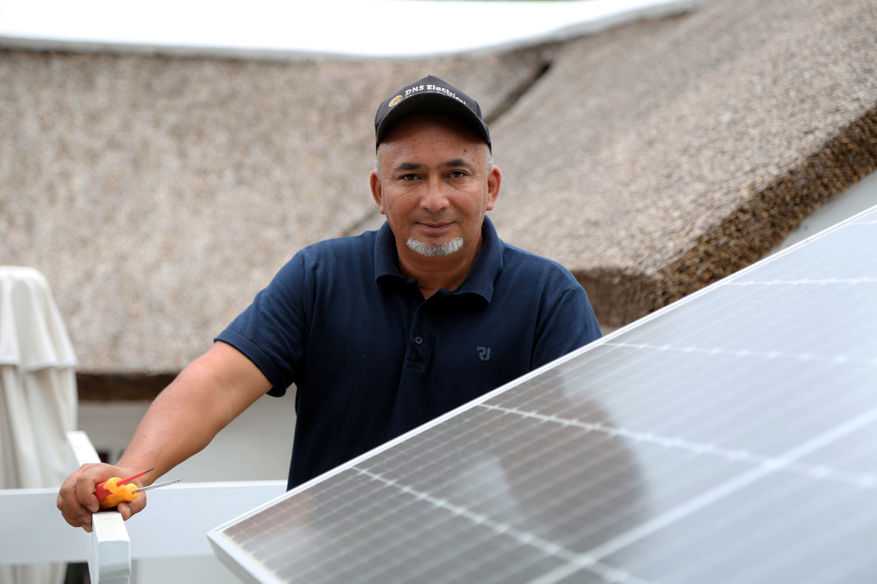 En man med keps håller i en skruvmejsel och står bredvid solpaneler. I bakgrunden finns en byggnad.