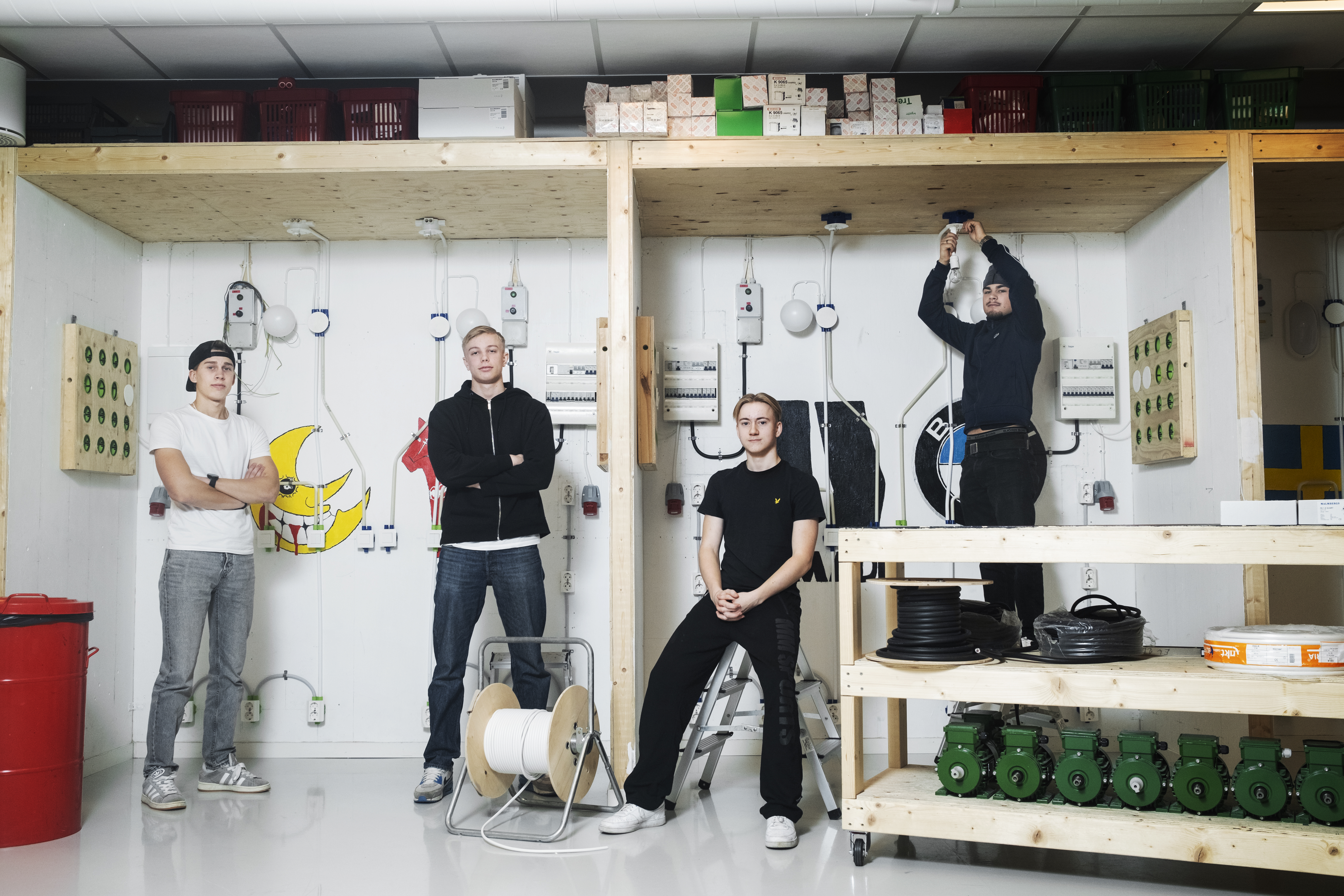 Fyra unga människor står och jobbar i en industriverkstad. I bakgrunden syns elutrustning, verktyg, kablar och trähyllor.