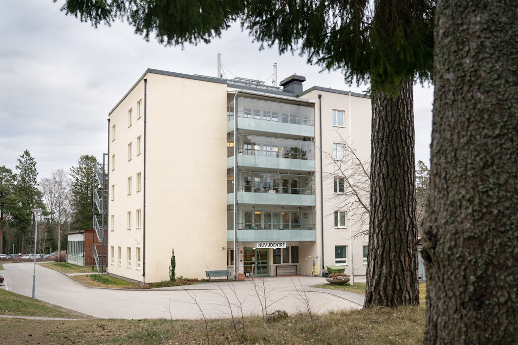 Ett fyravåningshus med en glasbalkong och en skylt där det står ”Huvudingång”, omgivet av träd och en asfalterad uppfart.