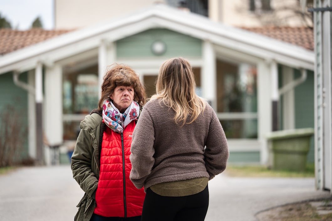 Två kvinnor står vända mot varandra och pratar utanför ett hus med vita kanter. Det är molnigt.