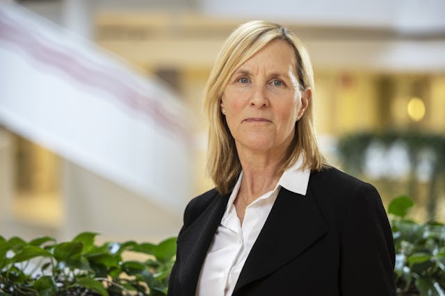 Lotta Wigen med blont, axellångt hår står inomhus framför en oskarp bakgrund med växter. Hon har på sig en svart kavaj och en vit skjorta.
