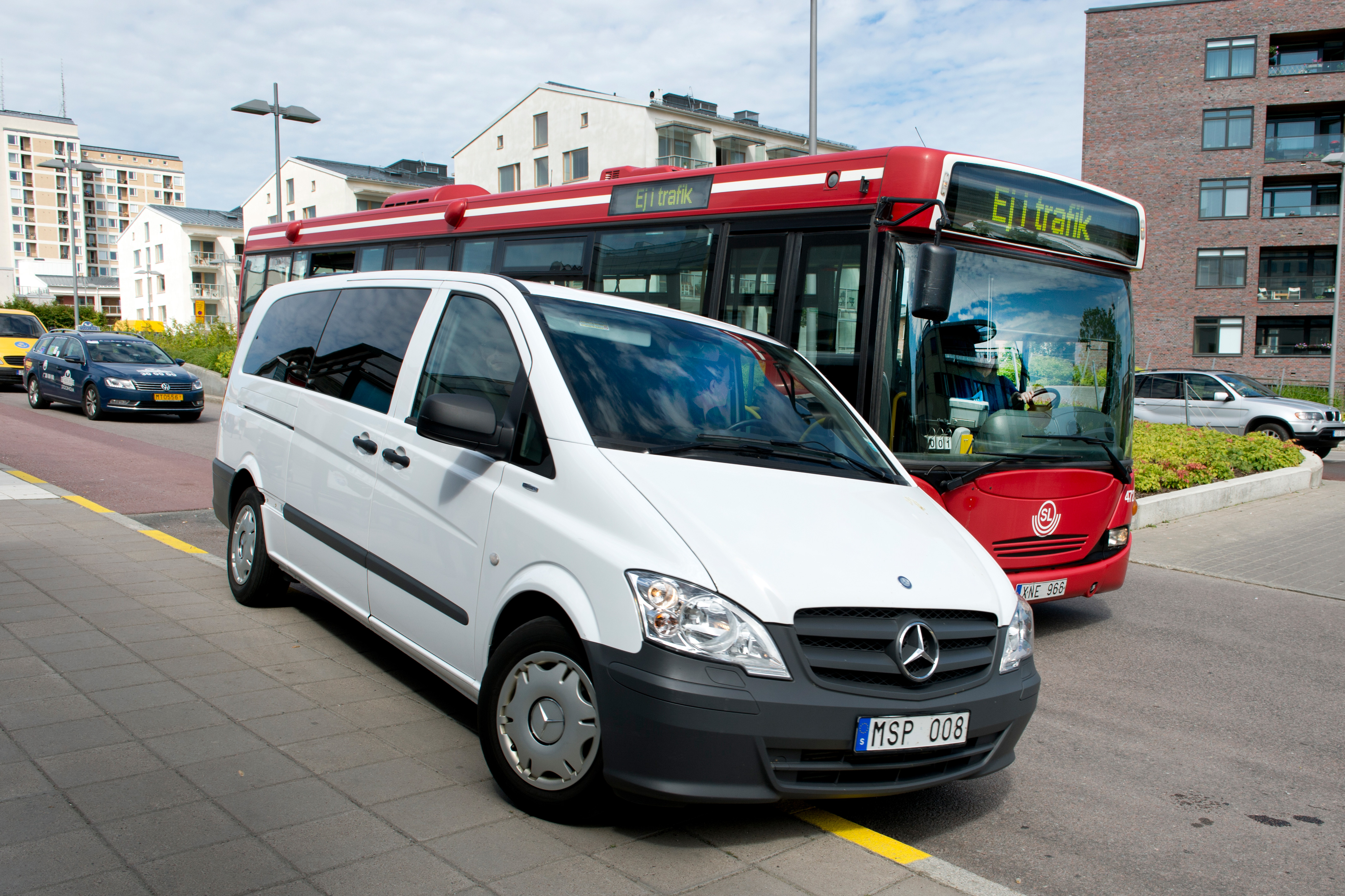 En Mercedes-Benz skåpbil står parkerad på trottoaren framför en stadsbuss på en gata i ett område med nya byggnader.