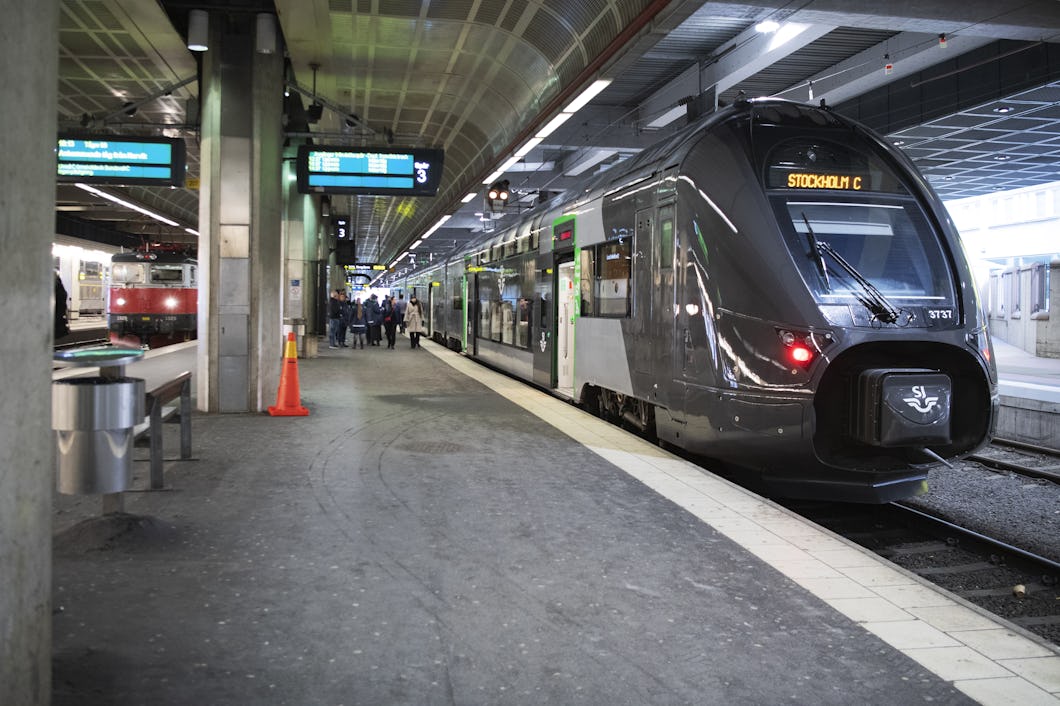 Ett tåg märkt "Stockholm C" står stilla vid en inomhusperrong. Passagerare och skyltar syns på plattformen.