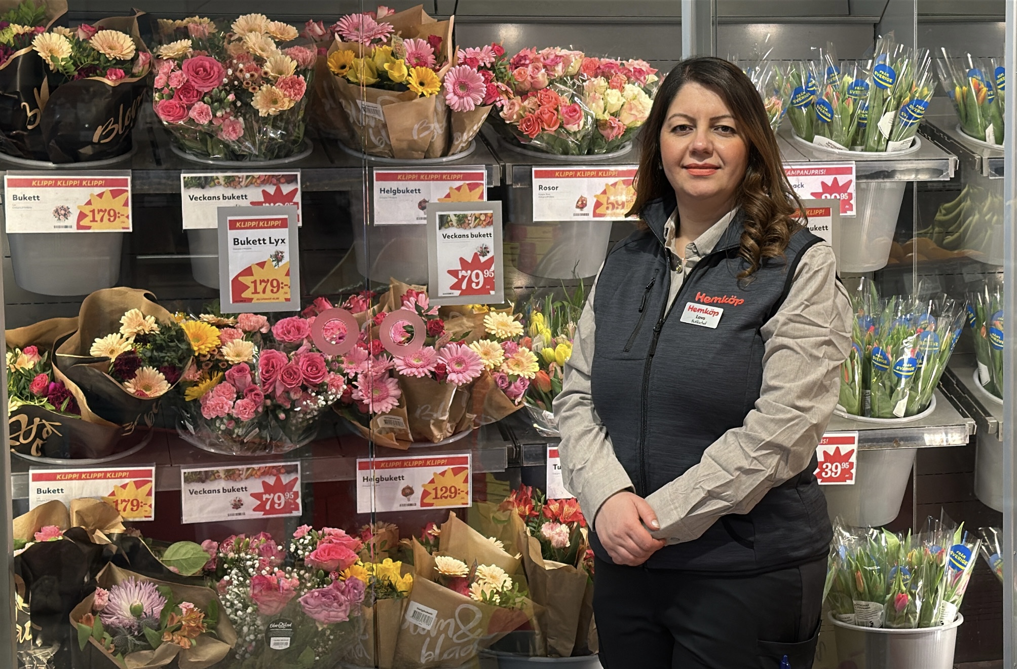 En butiksanställd står framför en blomsteravdelning med olika buketter. Prislappar och skyltar syns på hyllorna.