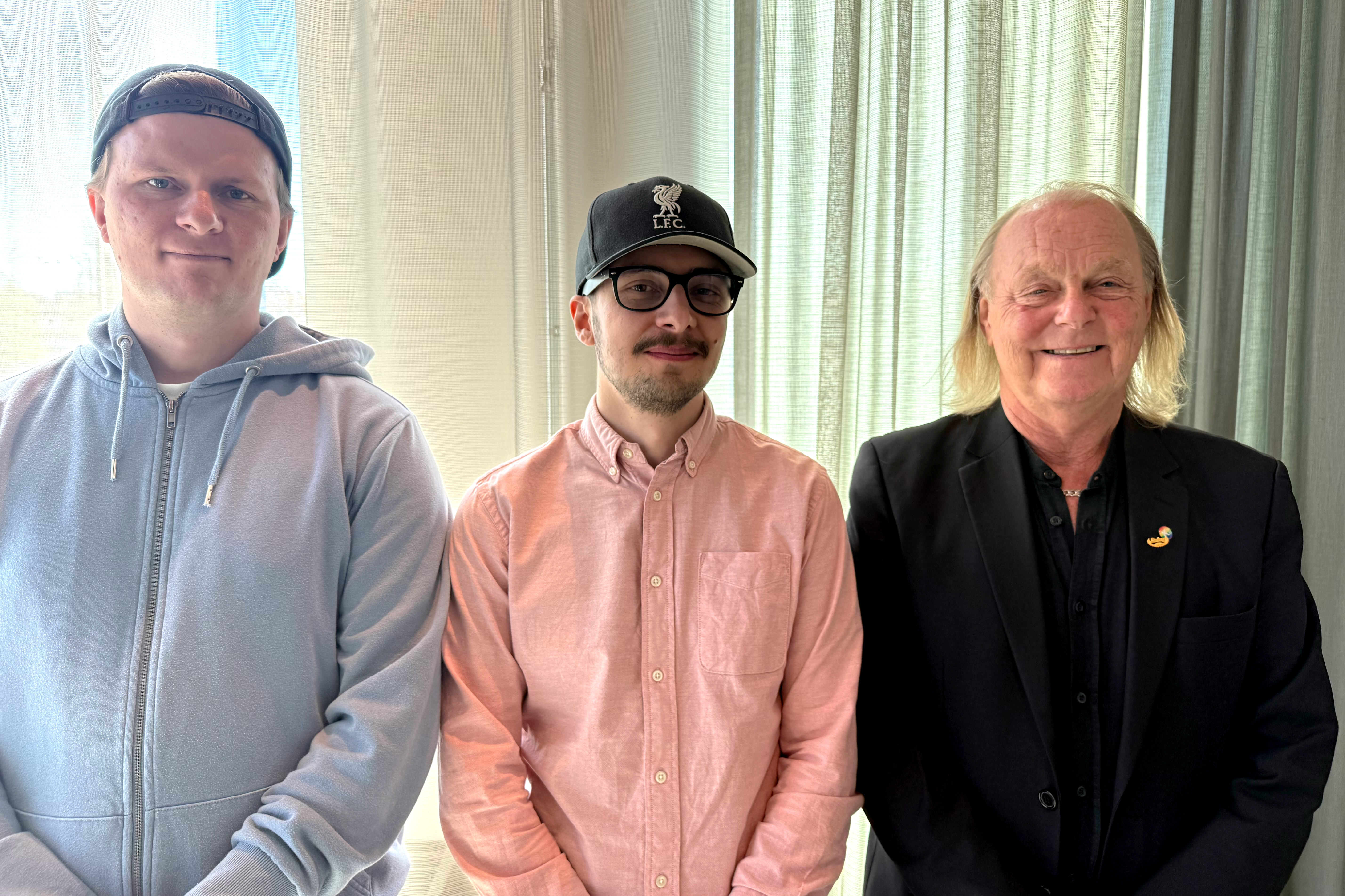Tre män står bredvid varandra inomhus framför genomskinliga gardiner och poserar för ett foto; två har på sig vardagskläder och en bär kostym.
