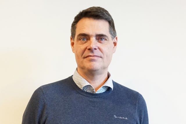 En man med kort brunt hår, iklädd en blå tröja över en skjortkrage, står mot en ljus bakgrund.