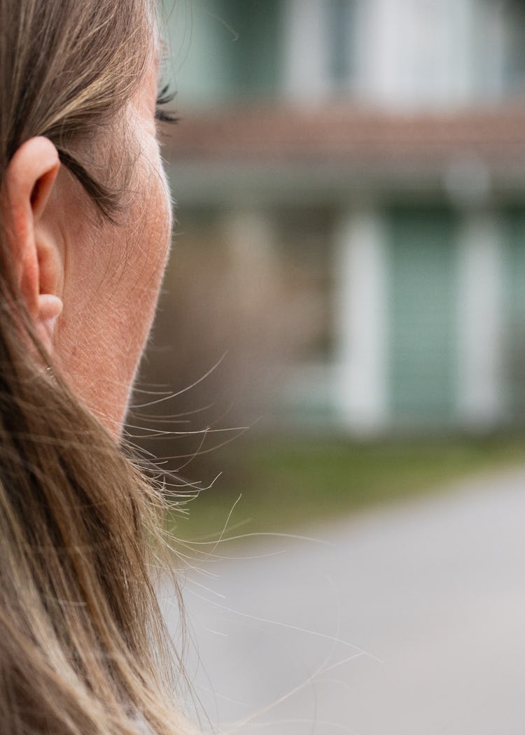 En anonym person med långt hår står utomhus, vänd mot en tvåvåningsbyggnad med veranda och vit foder, fotograferad bakifrån.