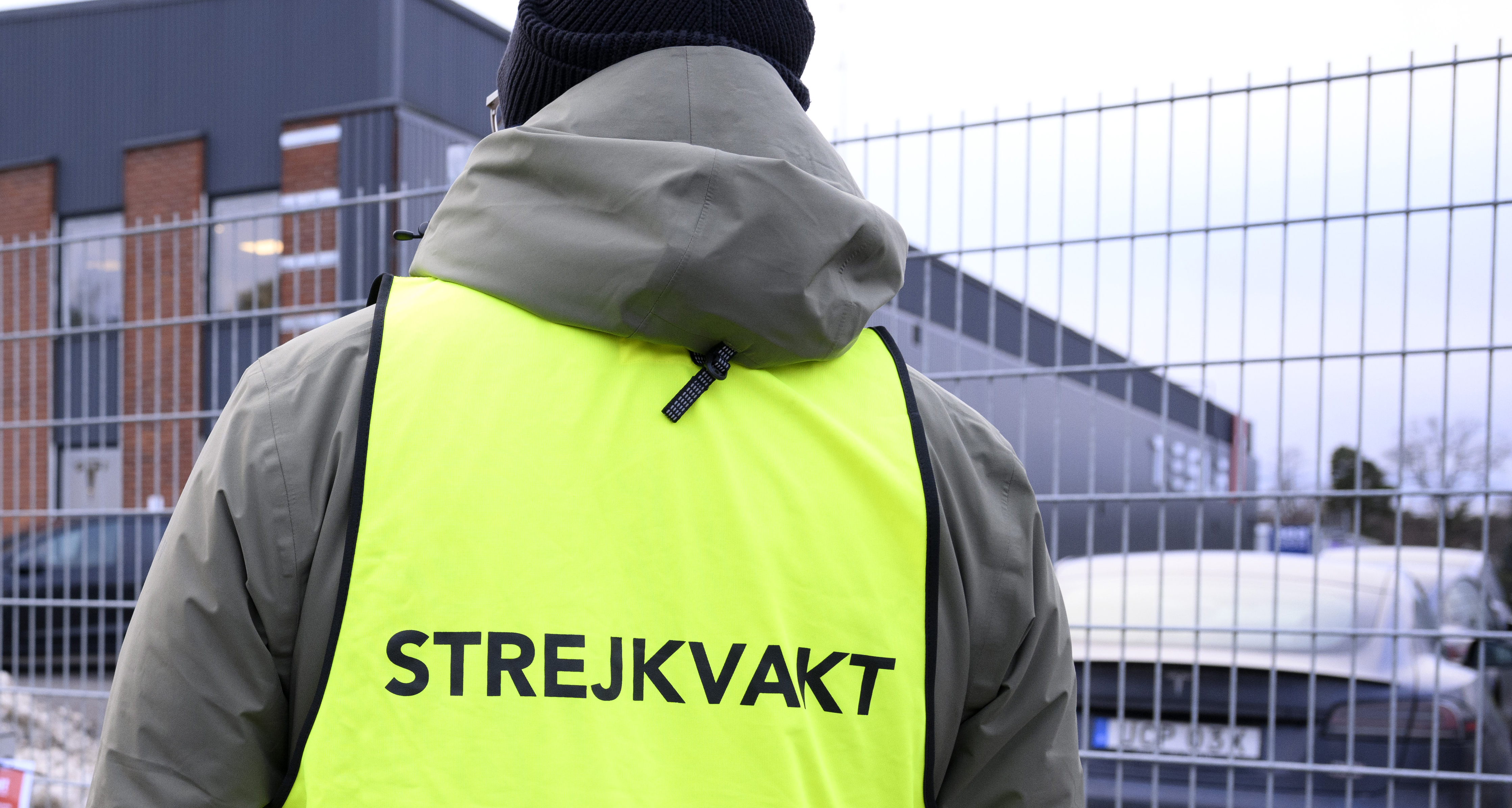 En person med en väst märkt "STREJKVAKT" står framför ett stängsel utanför en byggnad.