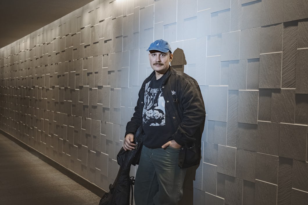 En man med mustasch har på sig en blå keps, en svart jacka och en mönstrad skjorta. Han står i en hall intill en vägg med geometriskt mönster.