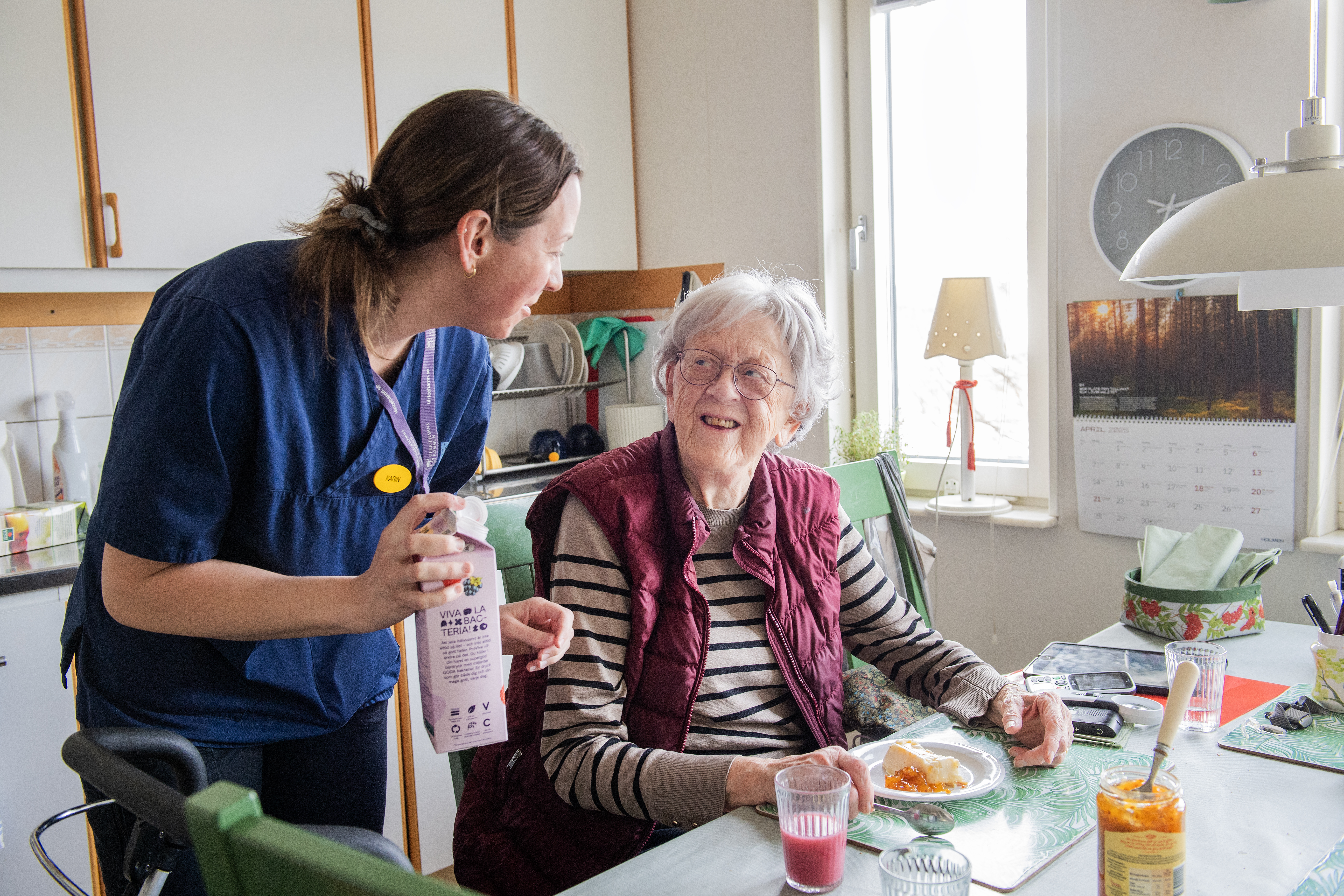 Karin Edelgripé räcker över en mjölkkartong till Inga Öjerbrand som sitter vid ett köksbord med mat och dryck.