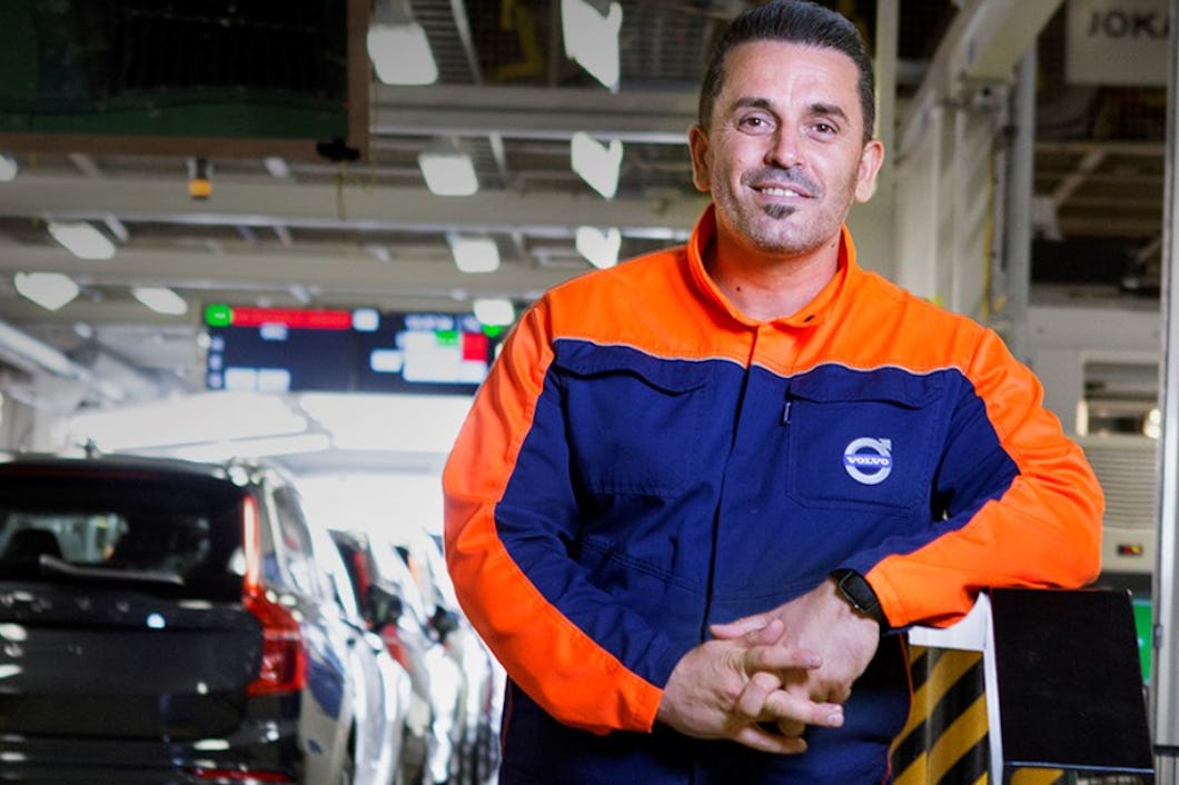 En man i Volvo-uniform står i en bilfabrik. Flera fordon och produktionsutrustning syns i bakgrunden.