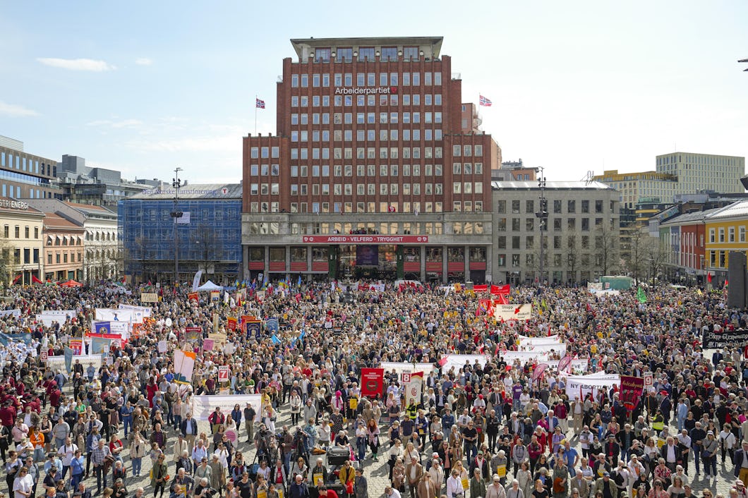 En folkmassa samlas på ett torg framför en byggnad av tegel med banderoller och skyltar för att delta i en demonstration.