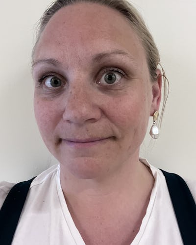 Sofia Stenman med ljust hår som är uppsatt och ljus hud står framför en ljus vägg. Hon har på sig en vit skjorta, mörka hängslen och ett pärlörhänge.