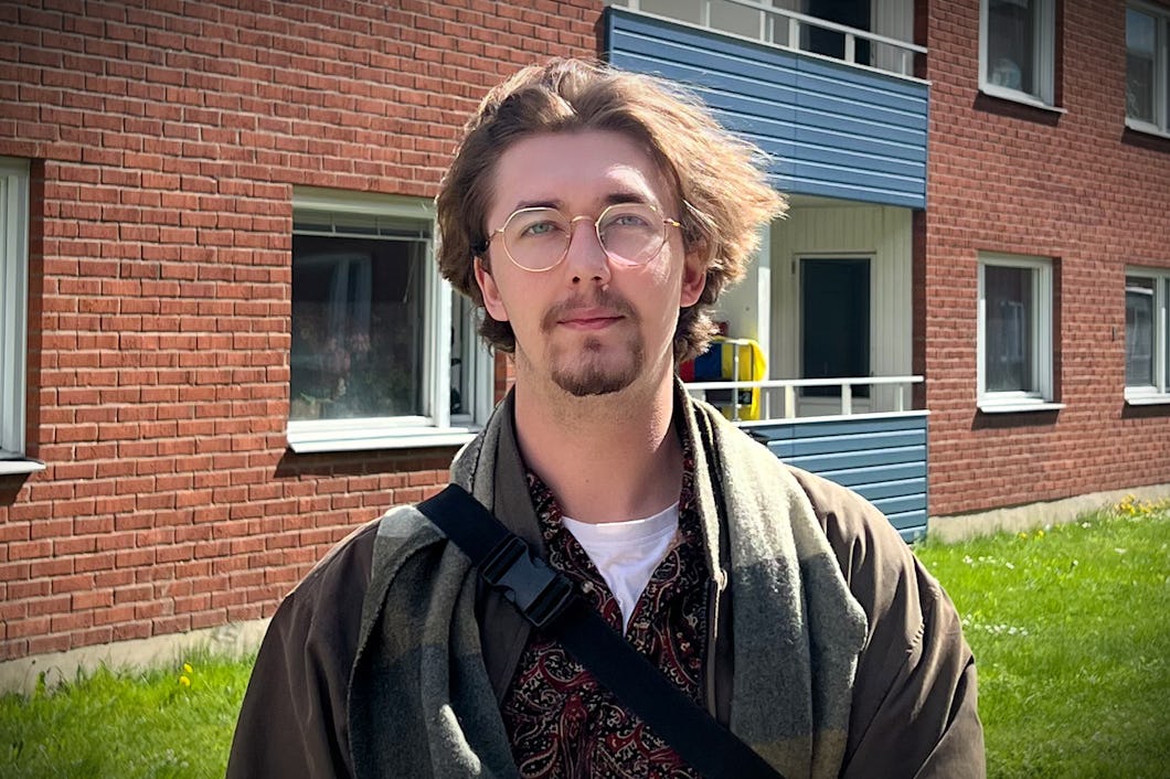 Loke Fors Olsson med glasögon och axellångt hår står utanför ett tegelhus och har på sig en halsduk och en axelväska.