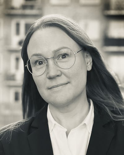 Cecilia Arklid med långt hår och glasögon, klädd i kavaj och vit skjorta, står framför en bakgrund med byggnader som inte syns tydligt.