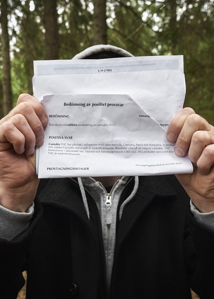 En person med kappa och huva håller upp och delvis döljer ett skrynkligt papper i en skog.