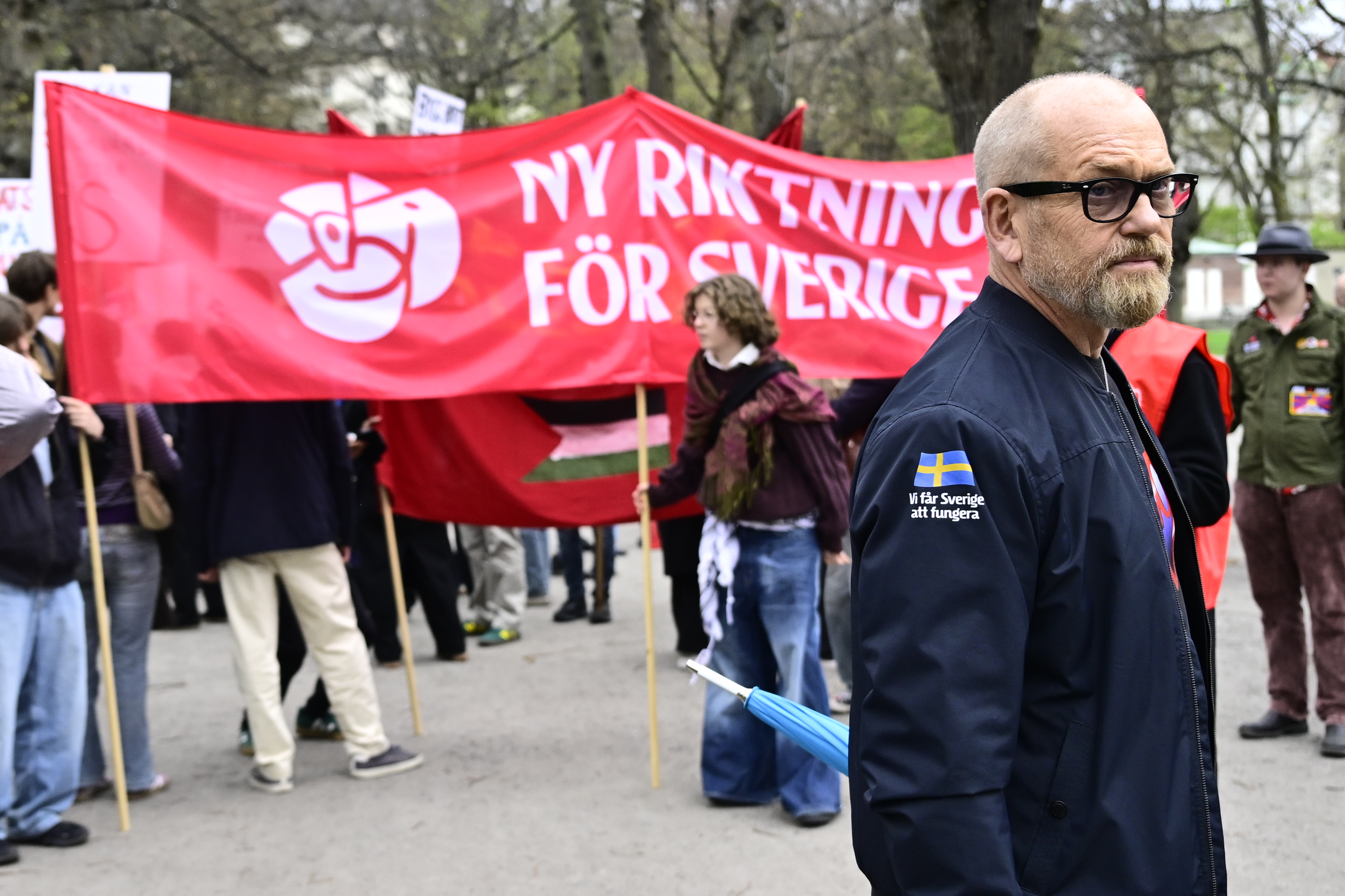 En man står framför en grupp och håller ett rött plakat med svensk text under en demonstration utomhus.