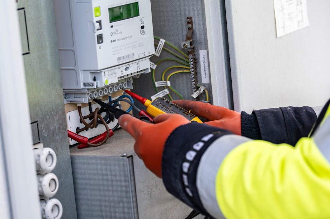 En person med skyddshandskar och varseljacka använder elektriska en multimeter för att testa ledningar i en styrpanel.