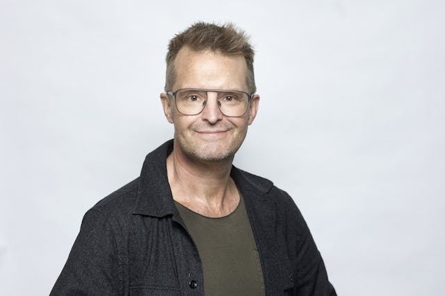 En man med kort hår och glasögon står framför en vit bakgrund. Han har på sig en mörk jacka och en grön skjorta. Mannen är vänd mot kameran och ler svagt.