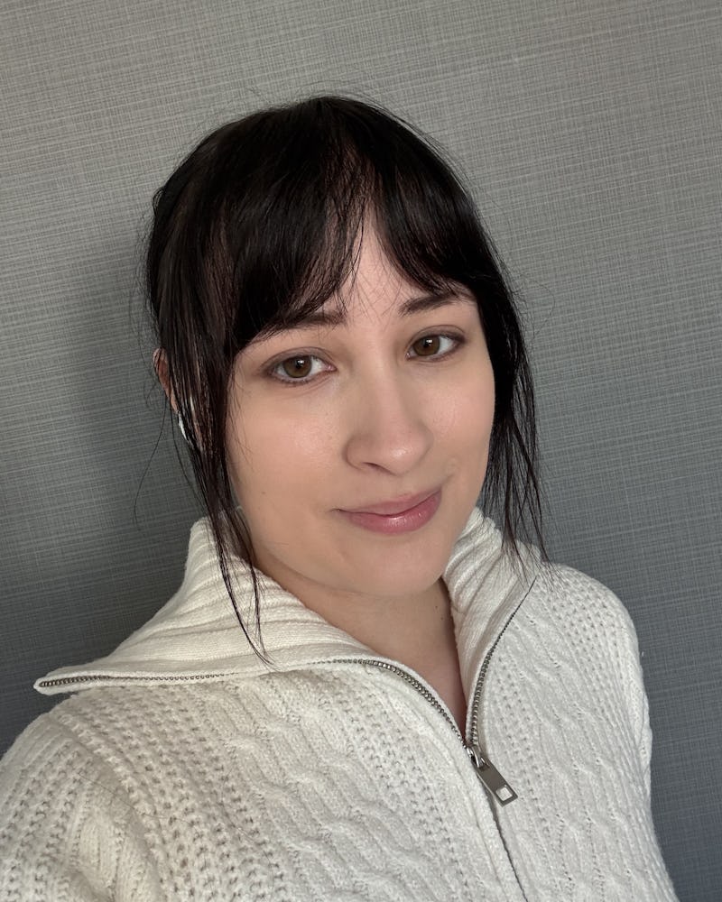 En person med rakt mörkt hår och bruna ögon har på sig en vit stickad tröja med dragkedja. Personen står framför en grå vägg och tittar in i kameran utan att le.