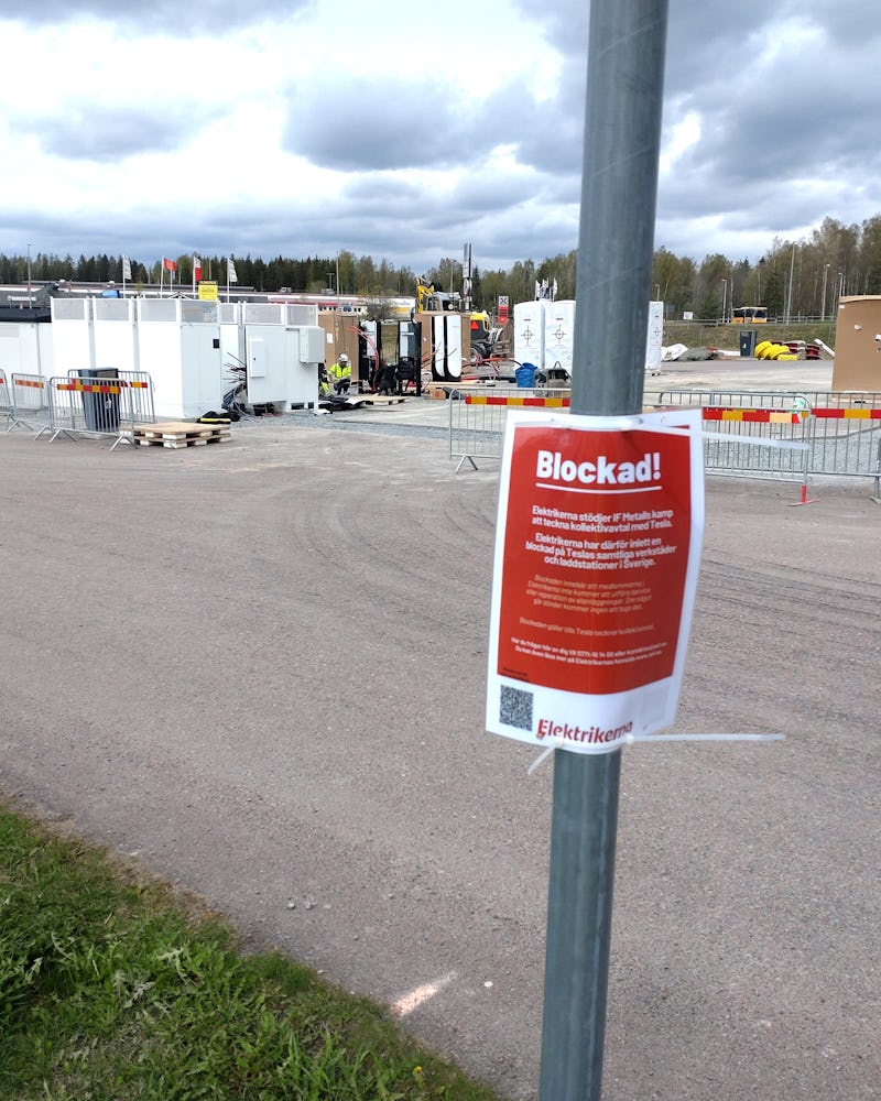 En röd skylt med texten "Blockad!" är fäst på en stolpe framför ett inhägnat byggande där det finns utrustning, baracker och arbetare i bakgrunden.
