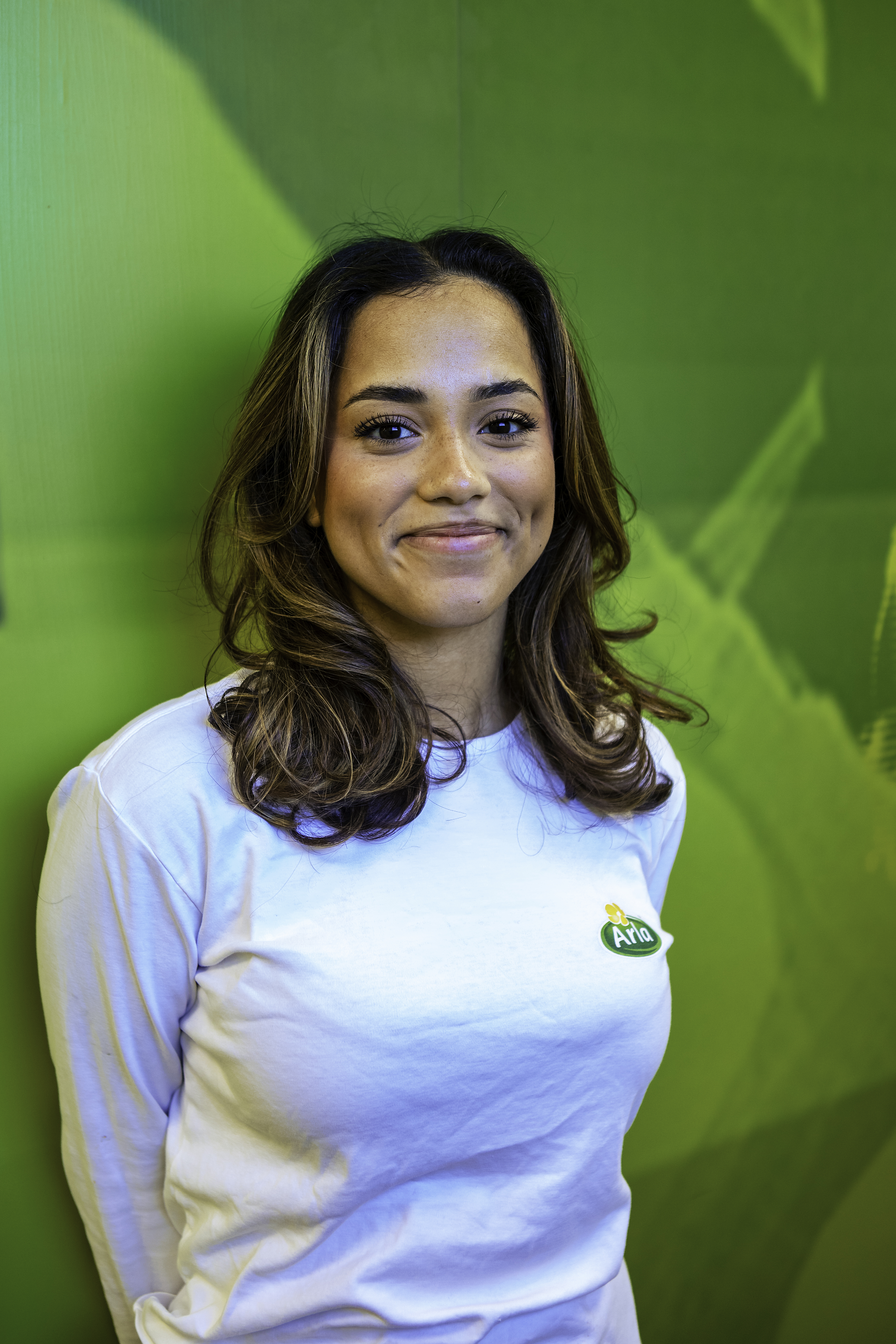 En kvinna med axellångt hår har på sig en vit långärmad tröja med "Arla"-logotyp och står framför en grön bakgrund.