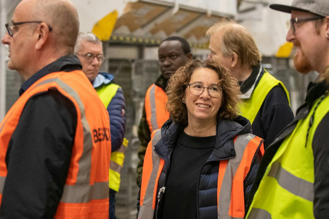 En grupp personer med säkerhetsvästar står tillsammans inomhus. Några av dem pratar med varandra. I bakgrunden syns hyllor och utrustning.