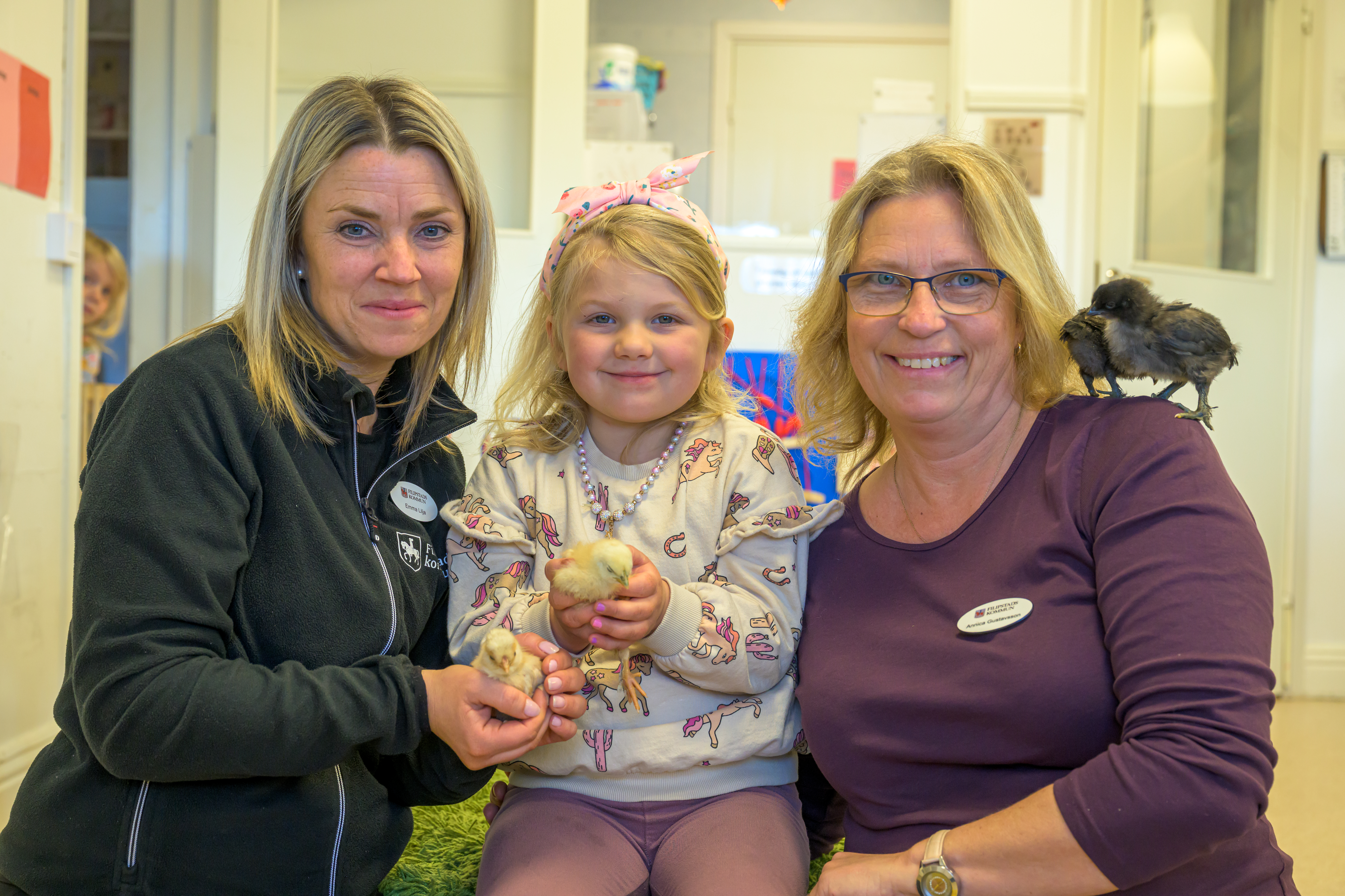 Tre personer sitter inomhus, Emma Lilja och Annica Gustavsson på varsin sida om Ebba som håller kycklingar; en av de vuxna har en svart kyckling på sin axel. Alla tittar mot kameran och ler.