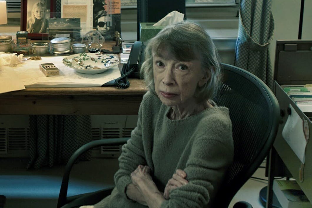 En äldre kvinna sitter vid ett skrivbord nära ett fönster. Stor text över bilden lyder: "Joan Didion: Anteckningar till John.