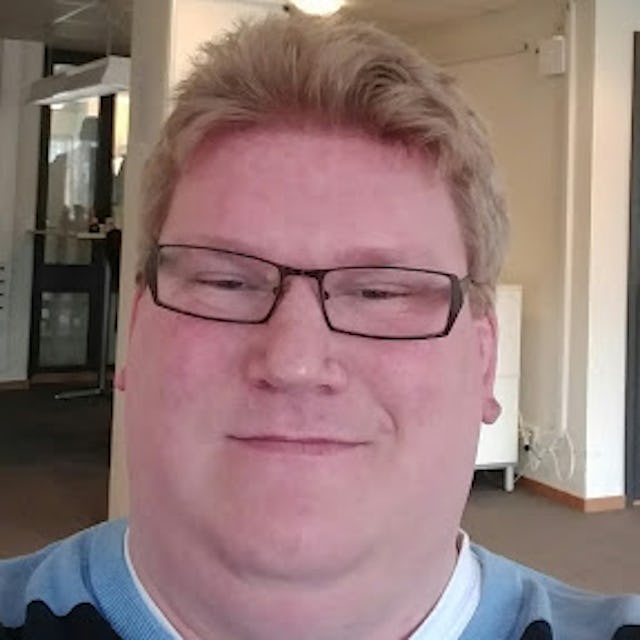 En person med kort blont hår och glasögon tar en selfie inomhus.
