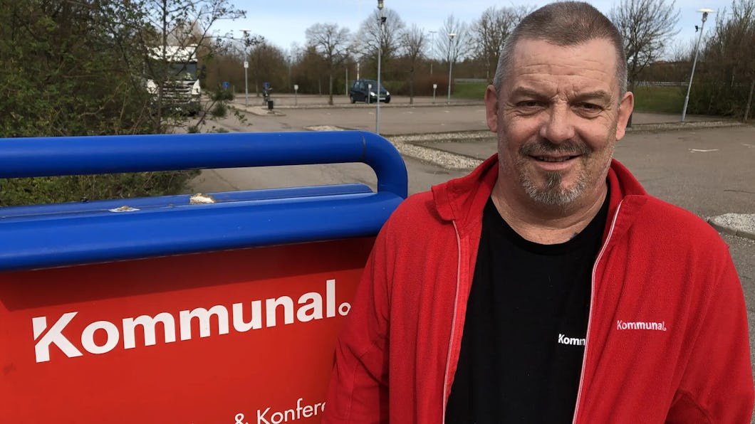 Mikael Olsson med en jacka från Kommunal står intill en skylt från Kommunal på en parkeringsplats utomhus, med träd och bilar i bakgrunden.