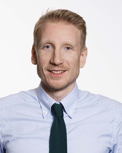 David Erixon med kort blont hår har på sig en ljusblå skjorta och en mörkgrön slips. Han ler mot kameran och står framför en ljus bakgrund.