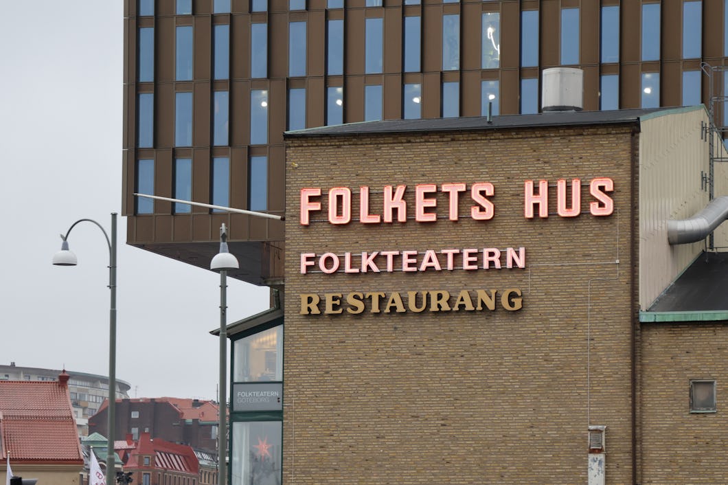 En tegelbyggnad med neonskyltar där det står "Folkets Hus," "Folkteatern" och "Restaurang," med både nya och äldre byggnader i bakgrunden.