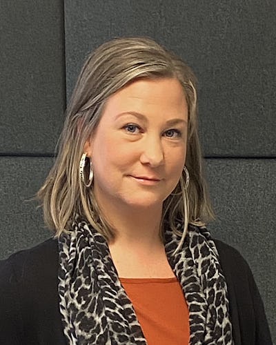 Annika Andersson med axellångt blont hår, som har ringörhängen, en svart topp, en orange skjorta och en sjal med leopardmönster står framför en mörk vadderad vägg.