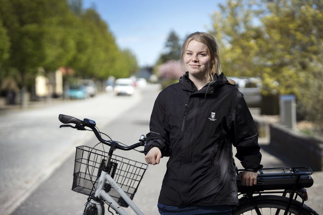 Isabel Weiland med en svart jacka står bredvid en cykel på en gata där det finns träd och bilar är parkerade längs trottoaren.