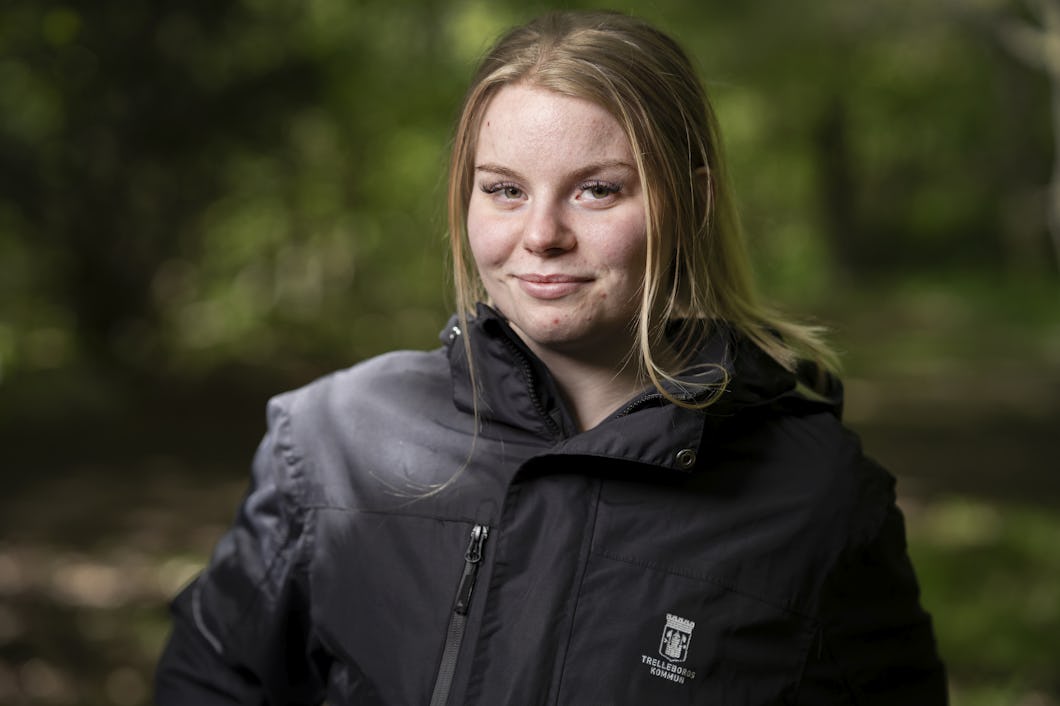 En kvinna med ljust hår står utomhus i ett skogsområde. Hon har på sig en mörk jacka märkt ”Trelleborgs kommun” och tittar mot kameran.