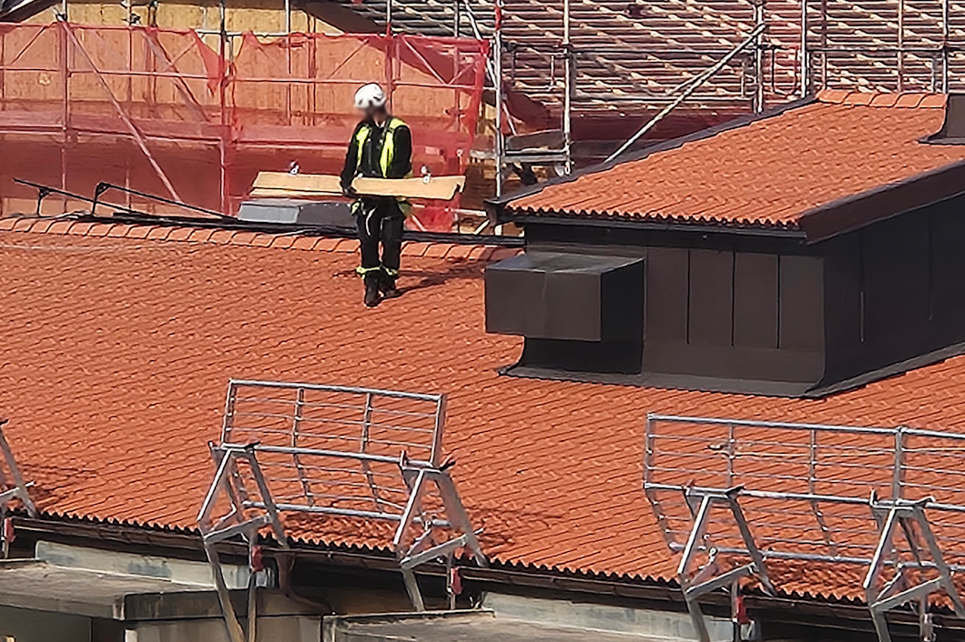 En byggarbetare med skyddsutrustning går på ett tak med tegelpannor och bär träplankor. Ställningar och byggmaterial syns i bakgrunden.