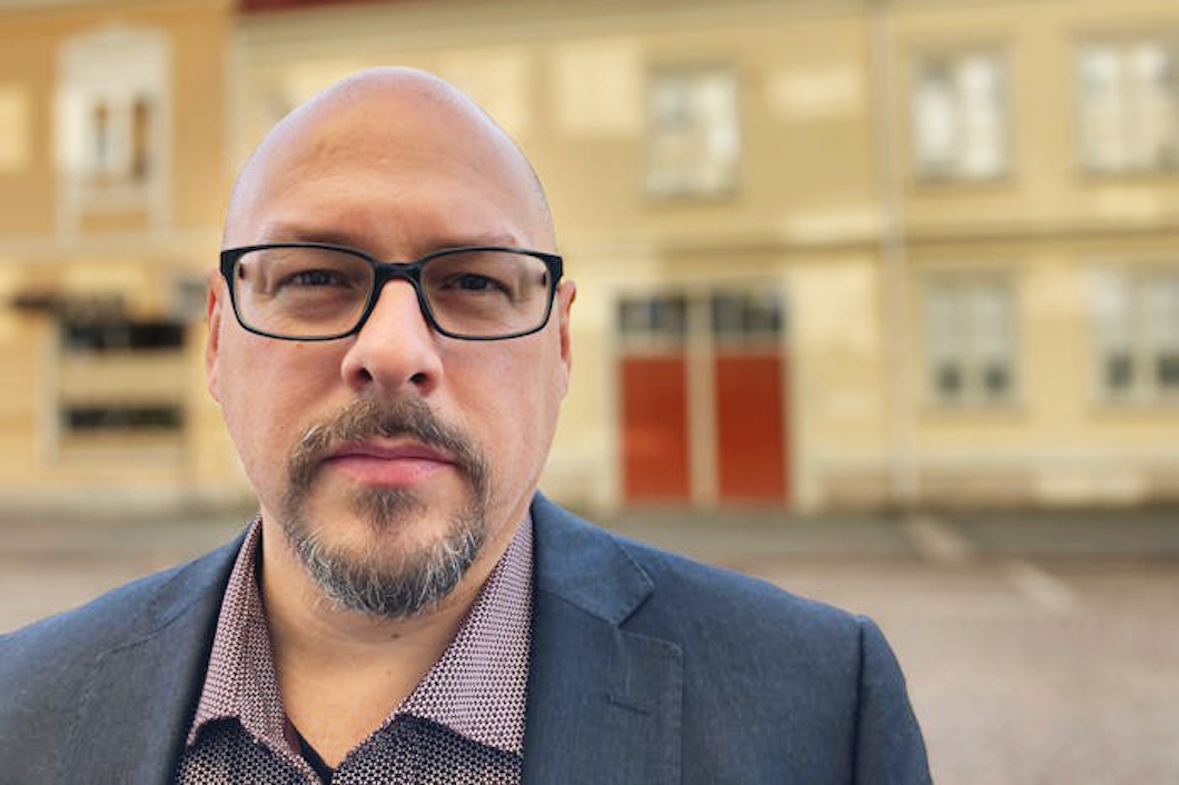 Anders Fennsjö med glasögon och skägg står utomhus utomhus en gul byggnad. Han har på sig kavaj och mönstrad skjorta.