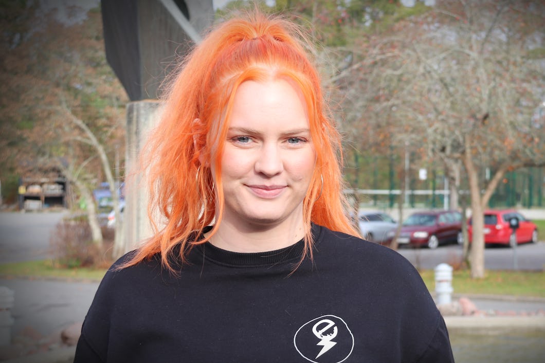 En person med orange hår och en svart sweatshirt står utomhus utomhus parkerade bilar och träd.
