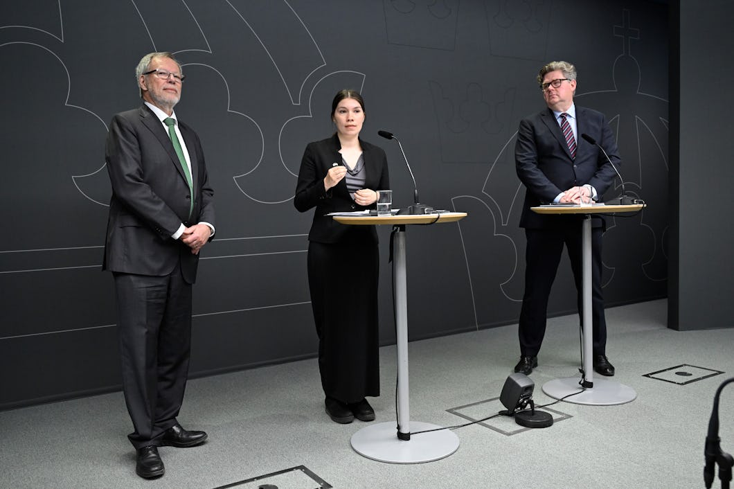 Tre personer står vid podier i ett rum för presskonferenser; två män i kostym står på varsin sida om en kvinna som tolkar till teckenspråk.