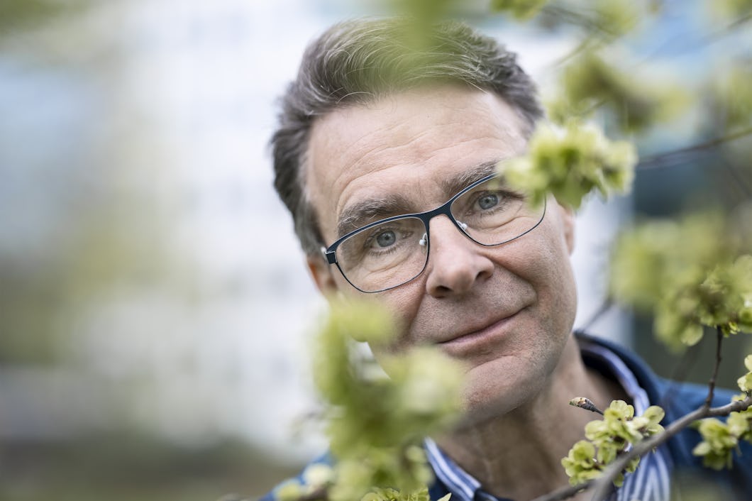 En man med glasögon tittar in i kameran genom grenar, med en suddig bakgrund.