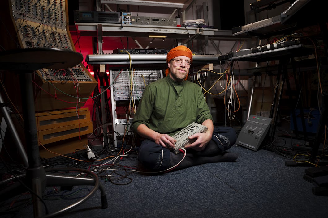 En person med huvudduk och grön skjorta sitter med korslagda ben på golvet, håller i en elektronisk apparat och är omgiven av syntar och ljudutrustning i en studio.