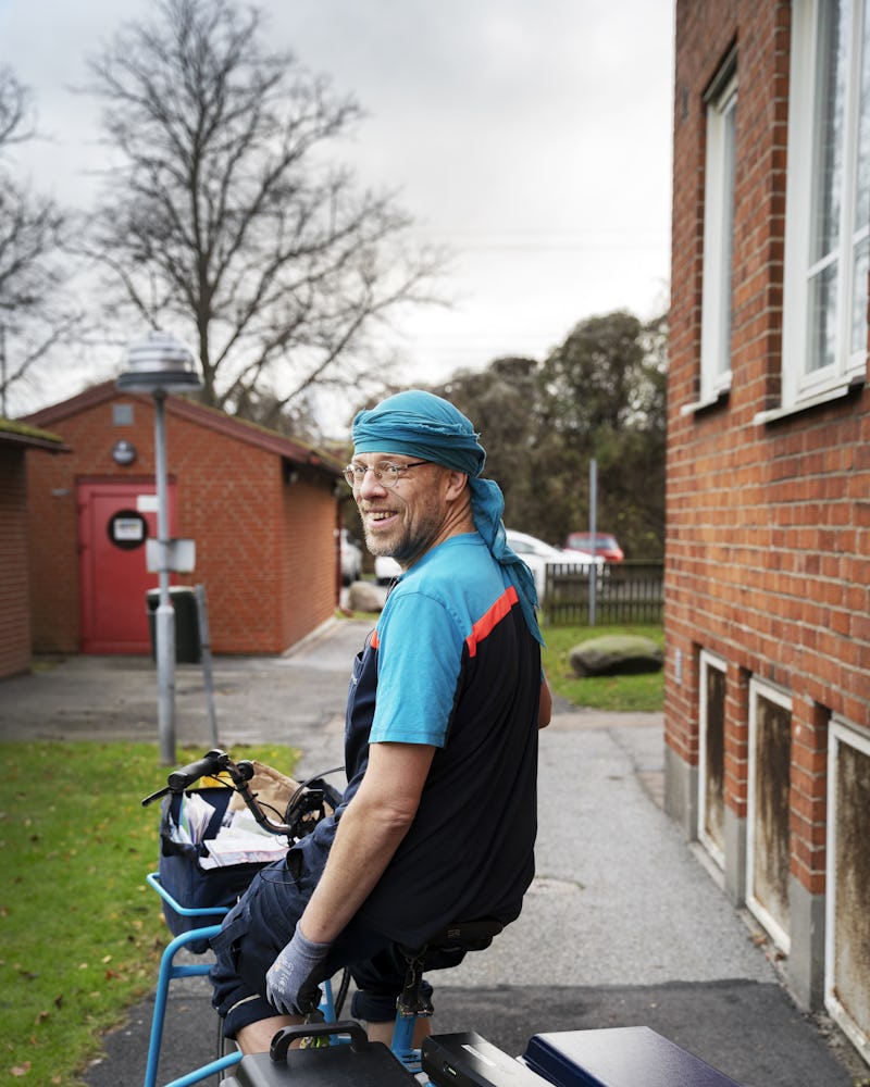 En man i uniform och huvudbonad sitter på medicinsk utrustning utanför en byggnad av tegel. Han tittar bakåt och ler.