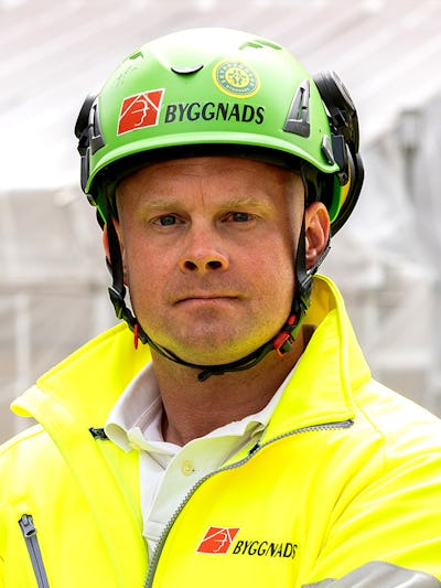 En person har på sig en gul jacka och en grön skyddshjälm med "Byggnads" logotyper och står i en utomhusarbetsmiljö.
