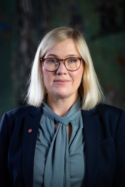 Sara Heelge Vikmång med rakt blont hår och glasögon har en blå kavaj över en grönblå blus och står framför en mörk, suddig bakgrund.