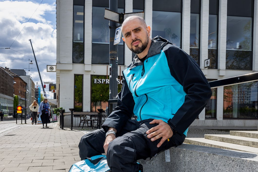 Ammar klädd i en turkos och svart jacka sitter på en betongbänk i ett stadsområde där det finns byggnader och människor i bakgrunden.