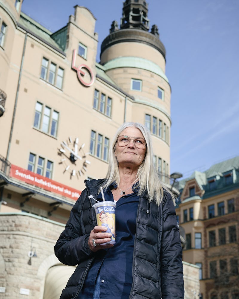 En kvinna med långt grått hår står utomhus och håller i en dryckesmugg. Hon befinner sig framför en byggnad med klocka och torn.