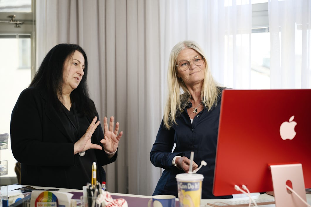 Två kvinnor står i ett kontor och pratar med varandra samtidigt som de tittar på en Apple-dator. En av dem gestikulerar med händerna.